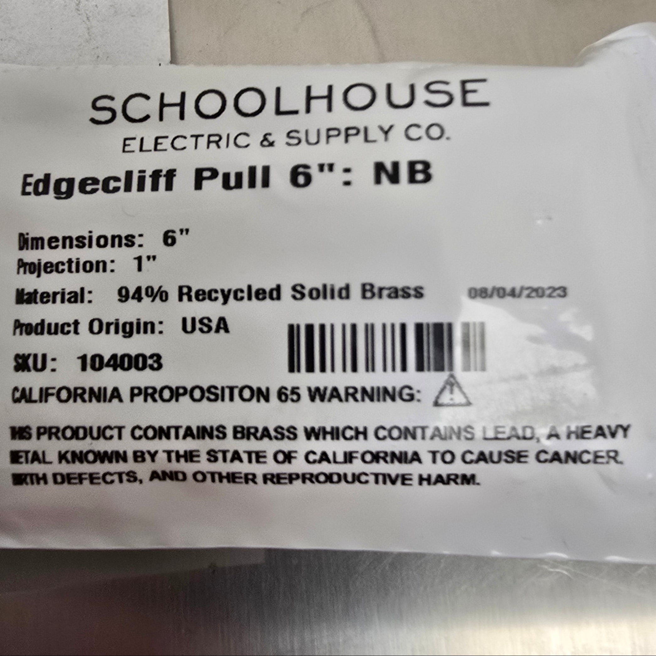 A white plastic-wrapped product label for Schoolhouse Electric & Supply Co. Edgecliff Pull 6" NB, showing printed text, a barcode, and a warning on a metallic surface.