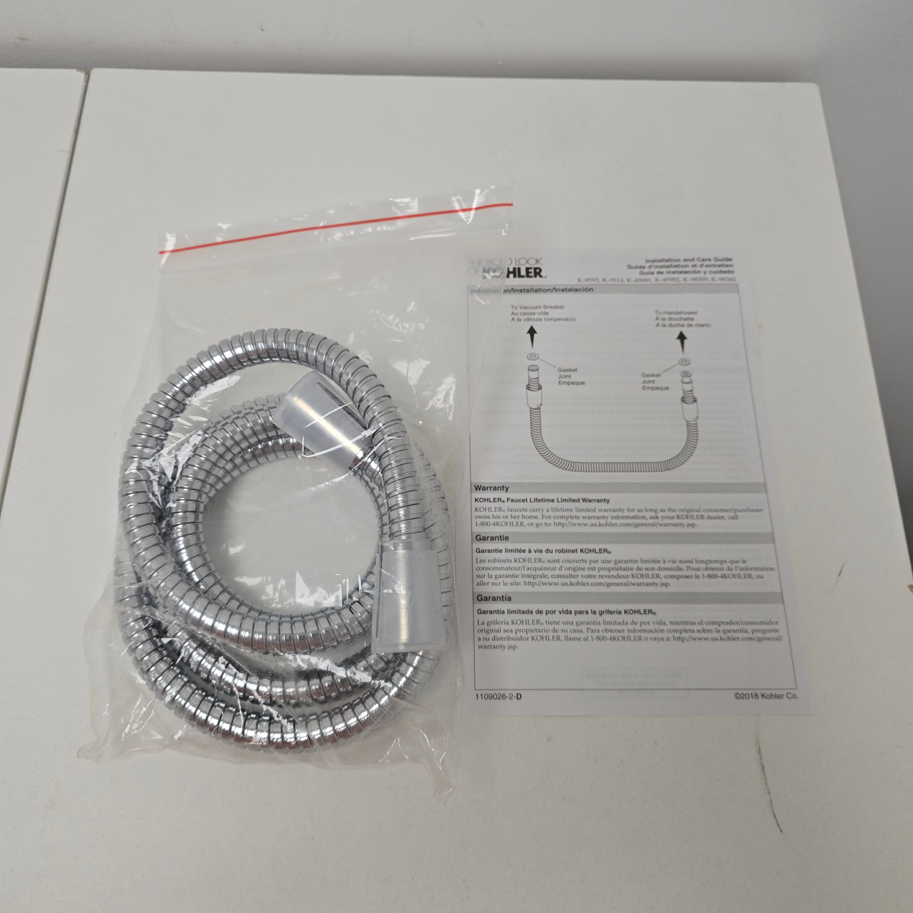 A coiled silver metal shower hose in a clear plastic bag, shown with a printed installation diagram card and a small red zip tie on a white surface.