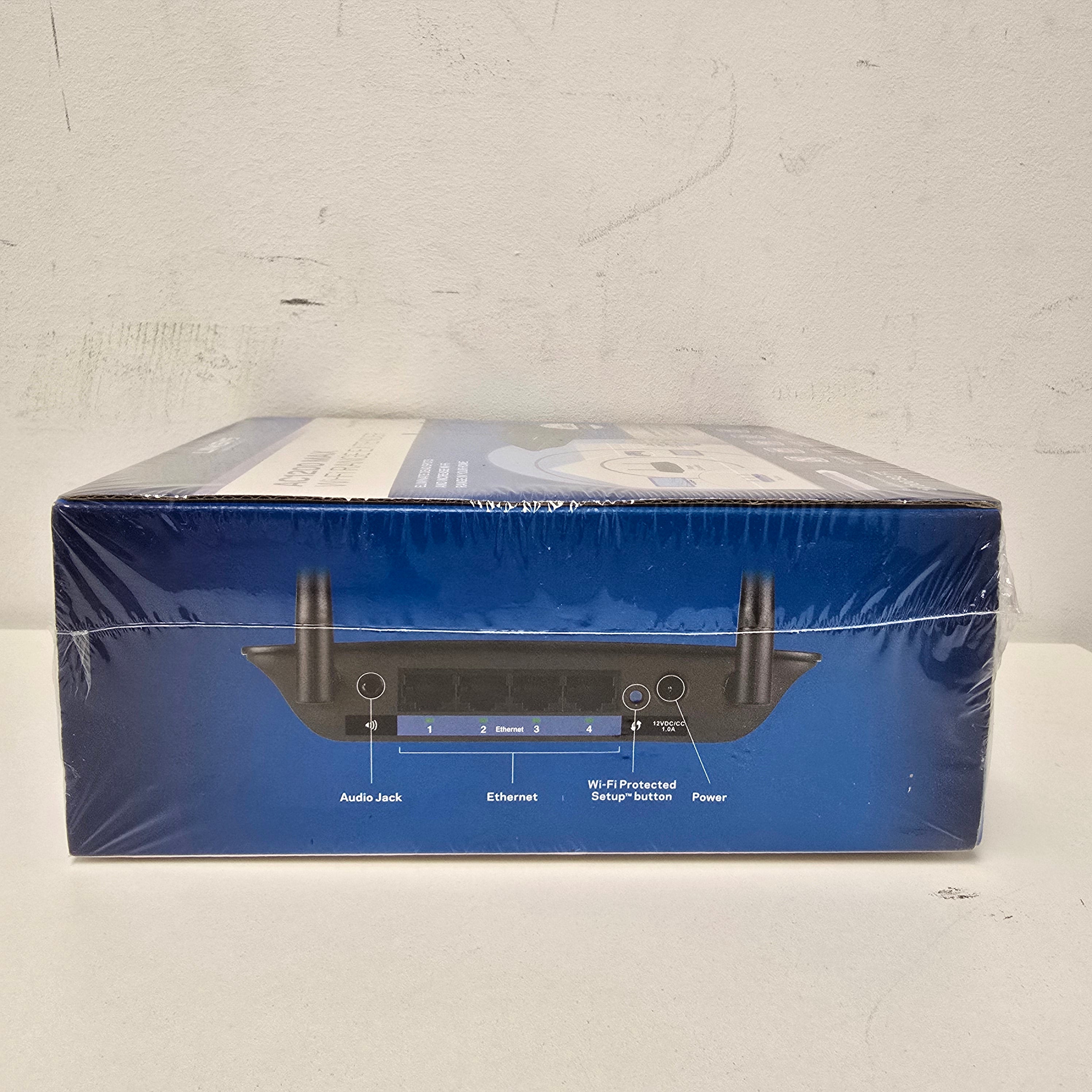 A blue retail box for a wireless router, wrapped in clear plastic, photographed on a white surface. The box shows a rear view of the router with labeled ports and antennas.